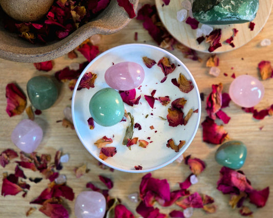 Top down view of the 8oz Heart Chakra Candle in a white tin with a polished Rose Quartz and Green Aventurine, surrounded by crystals and herbs on a wooden surface.