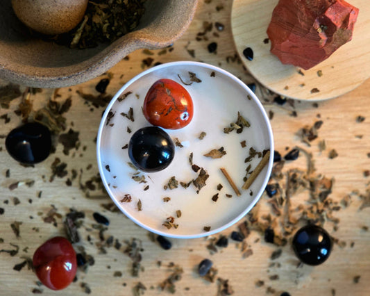 Top down view of the 8oz Root Chakra Candle in a white tin with a polished Red Jasper and Obsidian, surrounded by crystals and herbs on a wooden surface.