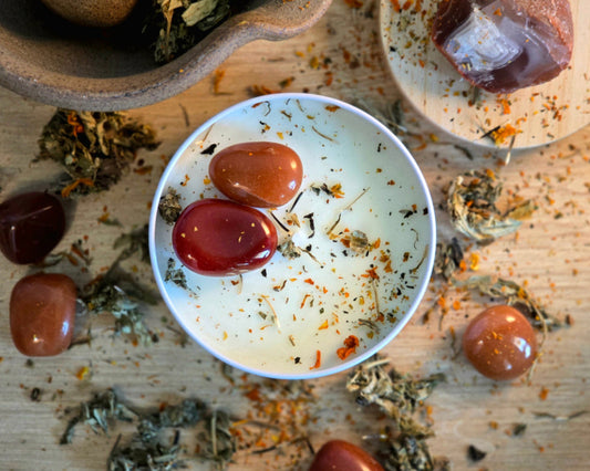 Top down view of the 8oz Sacral Chakra Candle in a white tin with a polished Carnelian and Red Aventurine, surrounded by crystals and herbs on a wooden surface.
