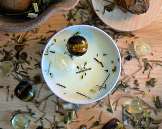 Top down view of the 8oz Solar Plexus Chakra Candle in a white tin with a polished Citrine and Tiger's Eye, surrounded by crystals and herbs on a wooden surface.