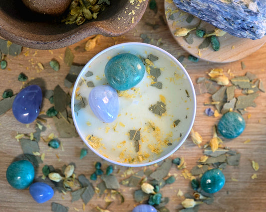 Top down view of the 8oz Throat Chakra Candle in a white tin with a polished Blue Lace Agate and Amazonite, surrounded by crystals and herbs on a wooden surface.