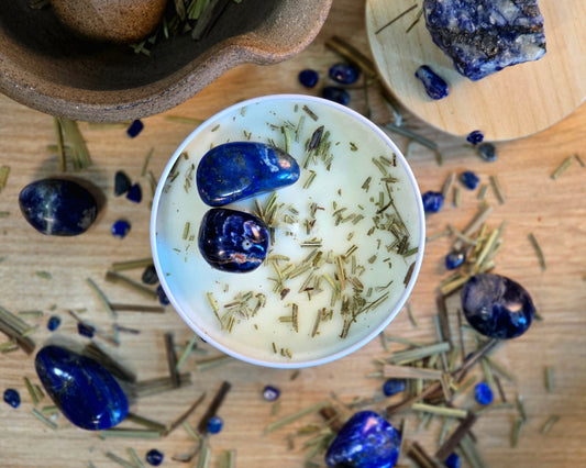 Top down view of the 8oz Third Eye Chakra Candle in a white tin with a polished Lapis Lazuli and Sodalite, surrounded by crystals and herbs on a wooden surface.