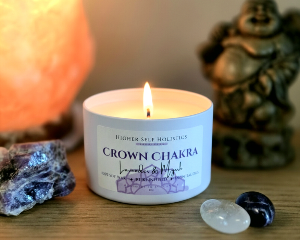 Crown Chakra Candle with a raw Amethyst Crystal, surrounded by crystals, with a salt lamp and buddha in the background, on a wooden surface.