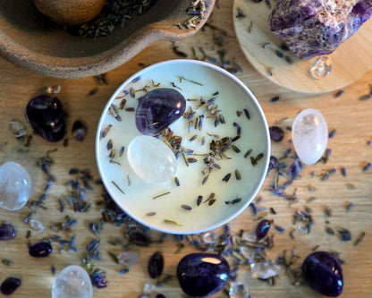 Top down view of the 8oz Crown Chakra Candle in a white tin with a polished Amethyst and Clear Quartz, surrounded by crystals and herbs on a wooden surface.