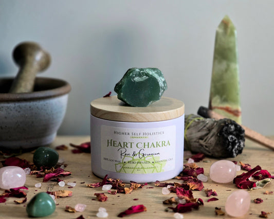 Heart Chakra Candle with a raw Green Aventurine Crystal on top of the lid, surrounded by crystals and herbs on a wooden surface.