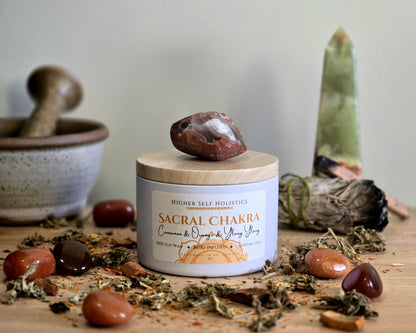Sacral Chakra Candle with a raw Carnelian Crystal on top of the lid, surrounded by crystals and herbs on a wooden surface.