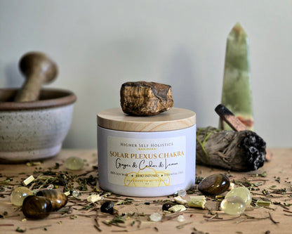 Solar Plexus Chakra Candle with a raw Tiger's Eye Crystal on top of the lid, surrounded by crystals and herbs on a wooden surface.