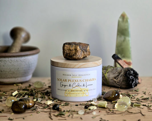 Solar Plexus Chakra Candle with a raw Tiger's Eye Crystal on top of the lid, surrounded by crystals and herbs on a wooden surface.