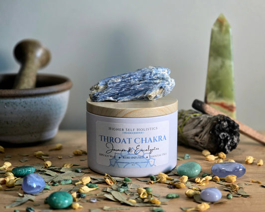 Throat Chakra Candle with a raw Kyanite Crystal on top of the lid, surrounded by crystals and herbs on a wooden surface.