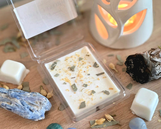 Throat Chakra Wax Melt with dried herbs in a clear container on a wooden surface with crystals, wax melt cubes, and a small wax melter.